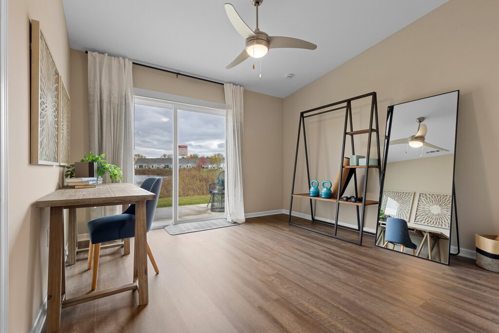 A modern room with wood flooring, a desk and chair by the window, open shelving with decor items, a large mirror, and sliding glass doors leading outside.