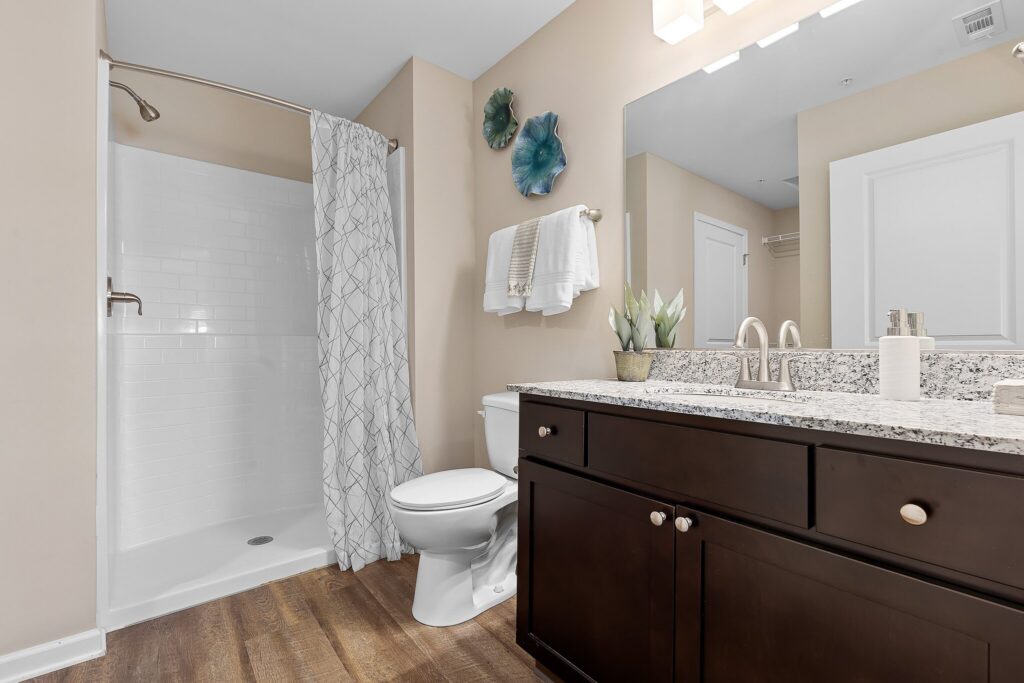 Modern bathroom with a glass-enclosed shower, patterned shower curtain, toilet, dark wood vanity with granite countertop, and wall decor above folded towels.