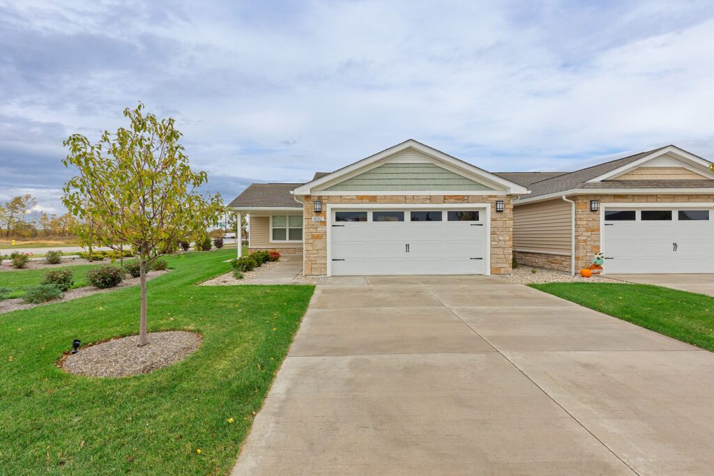 A driveway leads to a double garage attached to a single-story house with stone and siding exterior, landscaped lawn, and a small tree in front.