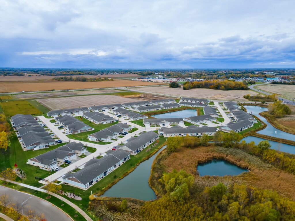 Aerial view of a suburban housing development surrounded by fields, water ponds, and patches of trees under a cloudy sky.