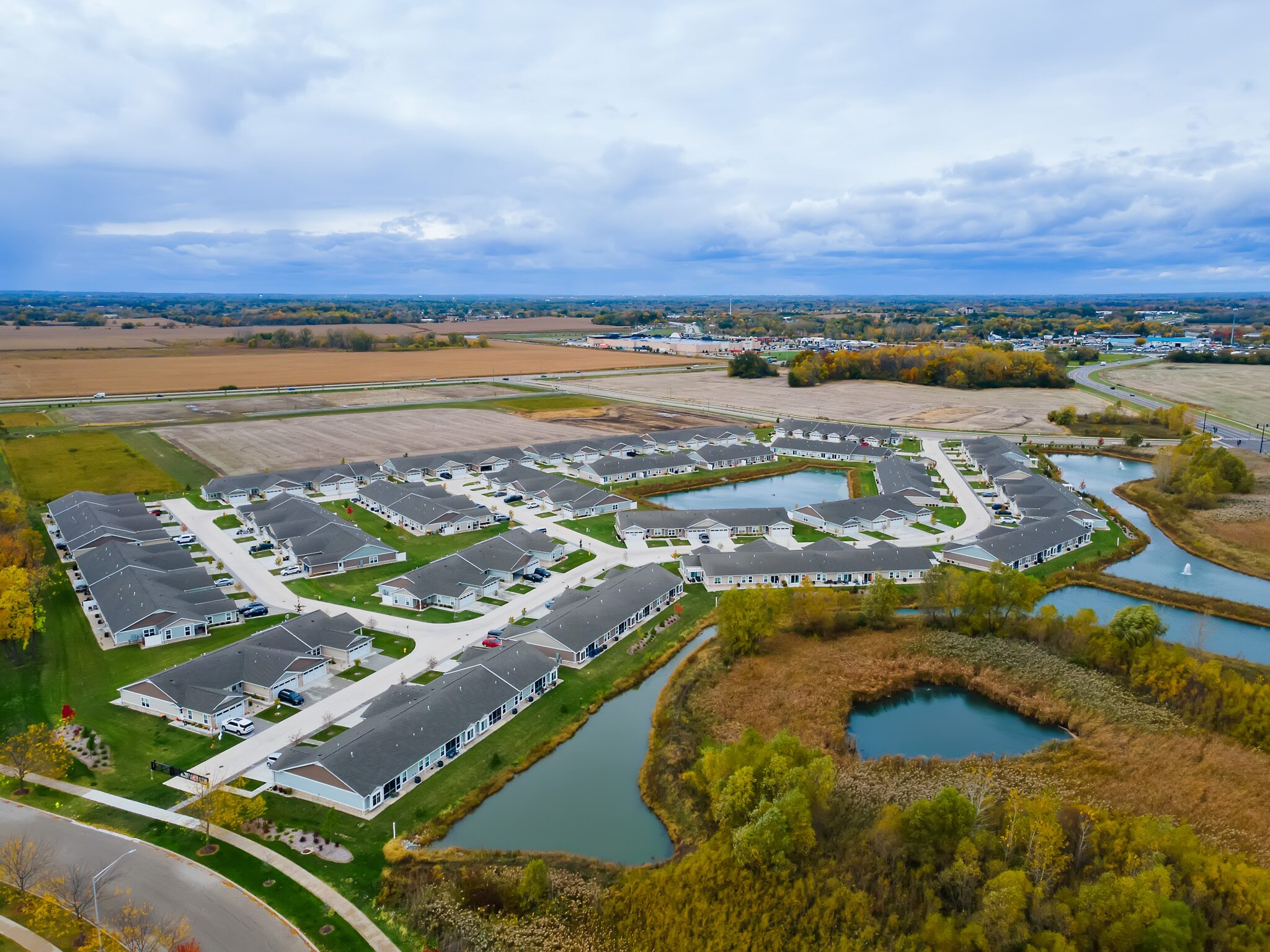 Aerial view of a suburban housing development surrounded by fields, water ponds, and patches of trees under a cloudy sky.