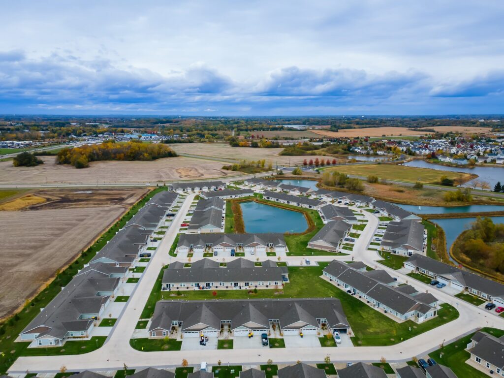 Aerial view of a suburban neighborhood with rows of houses, green lawns, small ponds, and surrounding farmland under a cloudy sky.