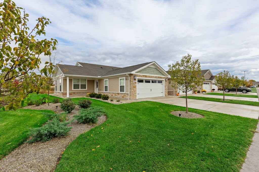 Single-story suburban house with a two-car garage, gray siding, and a well-maintained lawn with small trees and shrubs under a partly cloudy sky.