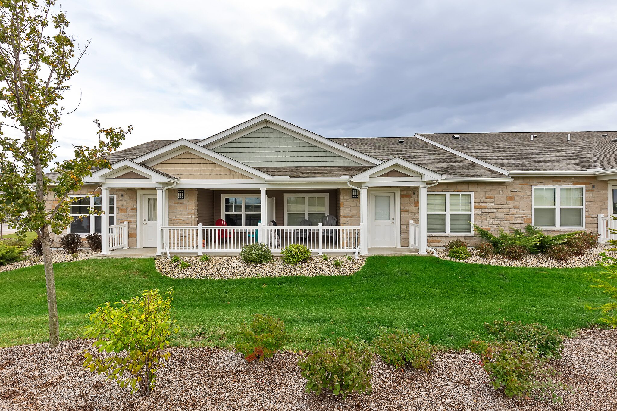Single-story duplex with a covered front porch, white railings, manicured lawn, shrubs, and cloudy sky overhead.