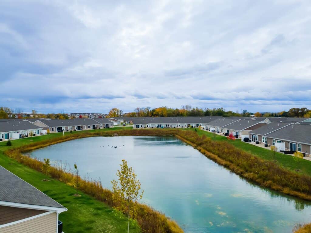 A small pond is surrounded by single-story homes and grassy areas, under a cloudy sky. Trees with autumn foliage are in the background.