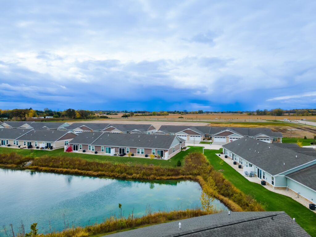 Rows of single-story houses with gray roofs border a small pond; fields and cloudy sky stretch in the background.