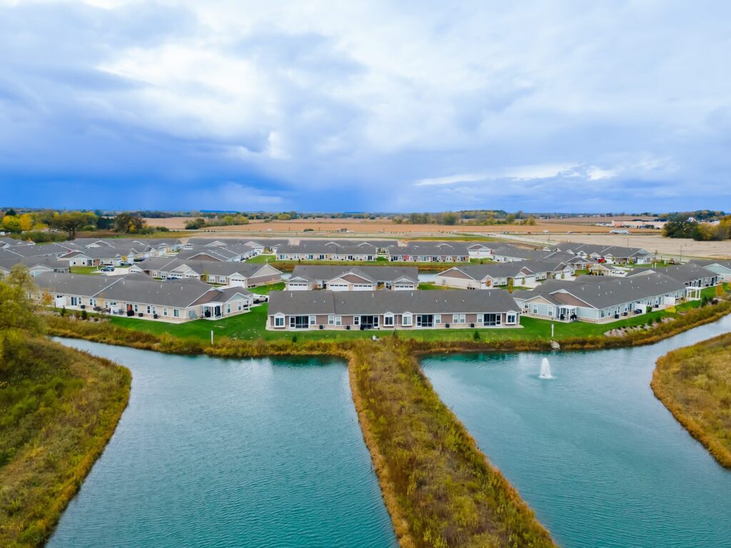 Aerial view of a suburban housing development surrounded by ponds and grassy areas, with farmland and cloudy sky in the background.