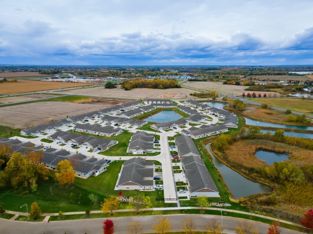 Aerial view of a suburban neighborhood with single-story houses, curved streets, ponds, and surrounding fields under a cloudy sky.