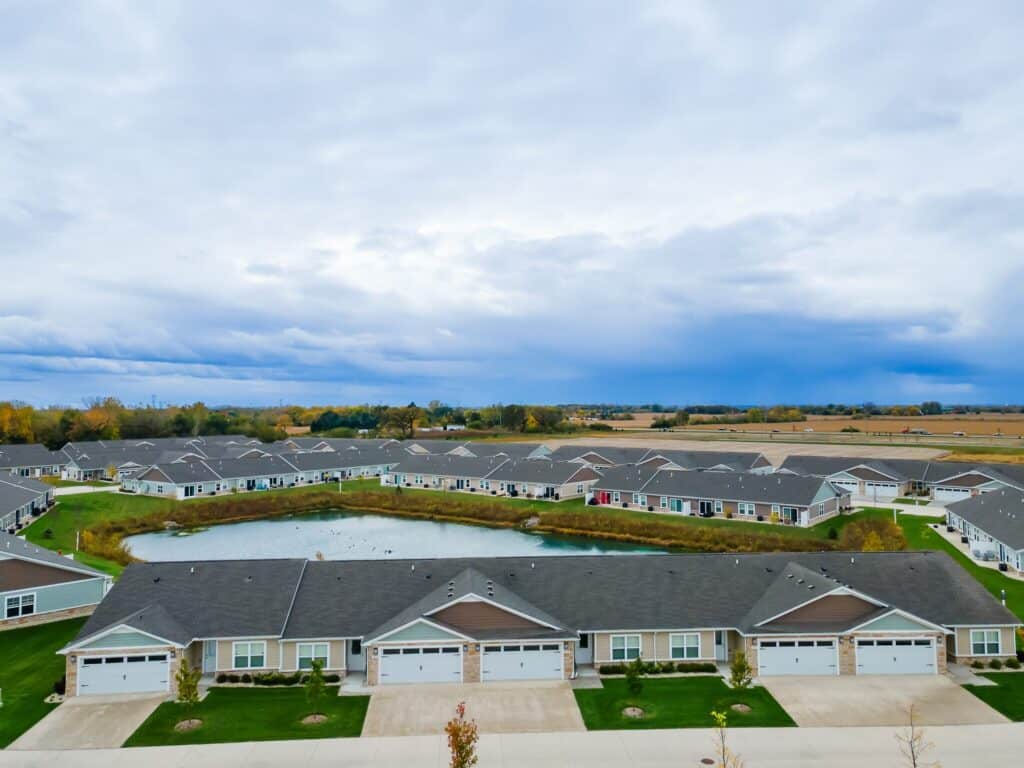 A suburban neighborhood with rows of single-story houses, each with a driveway and garage, surrounds a small pond under a cloudy sky.