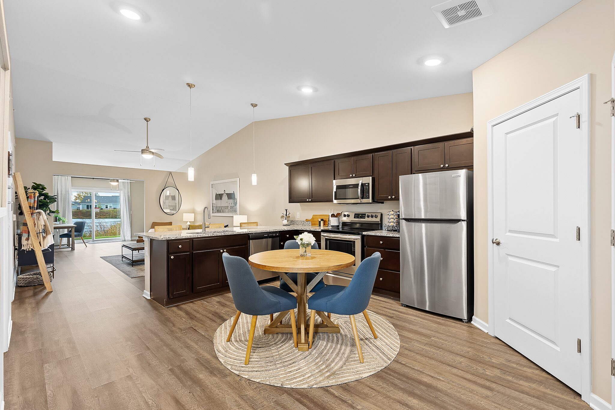 Modern kitchen and dining area with dark wood cabinets, stainless steel appliances, a round table with four blue chairs, and a living room visible in the background.