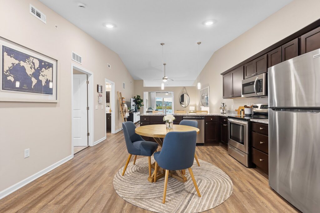 Modern kitchen and dining area with dark cabinets, stainless steel appliances, round wooden table with four blue chairs, and light wood flooring in open-plan space.