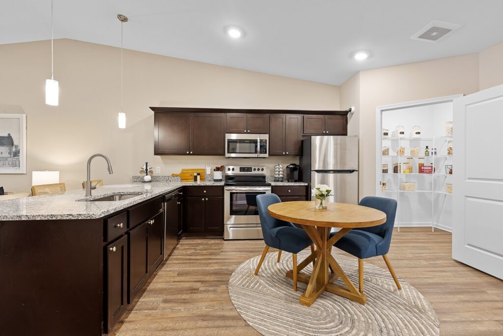 Modern kitchen with dark cabinets, stainless steel appliances, a round dining table with two blue chairs, and an open pantry with white shelves.