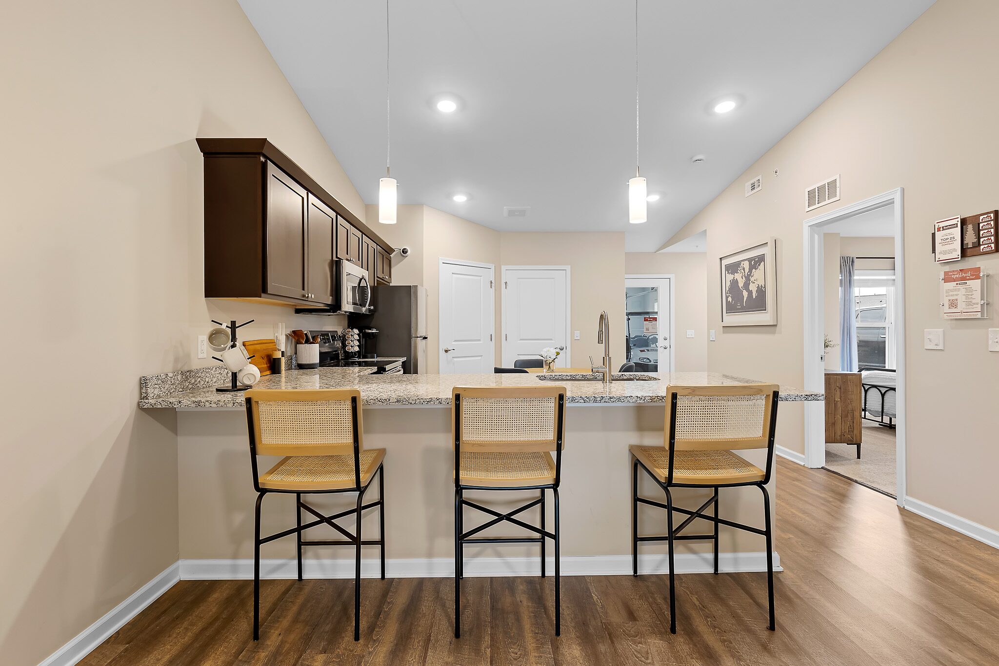 A modern kitchen with a granite island, three wooden bar stools, stainless steel appliances, and pendant lights, featuring beige walls and wood flooring.