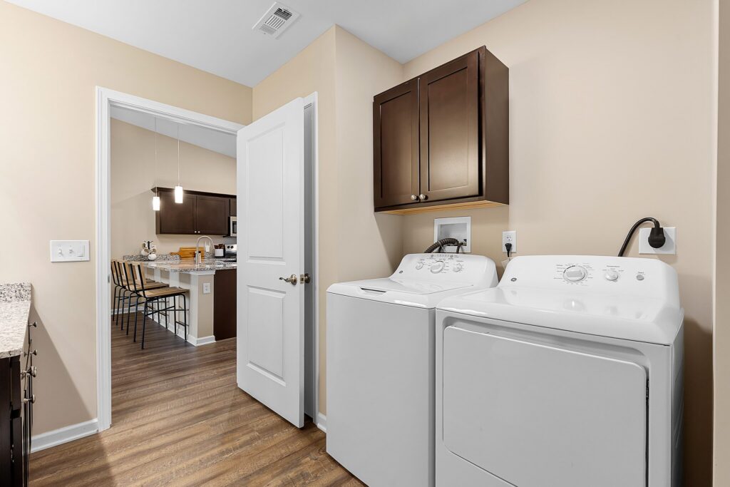 Laundry room with a white washing machine and dryer, dark wood cabinets above, and a view into a kitchen and dining area with wooden flooring.