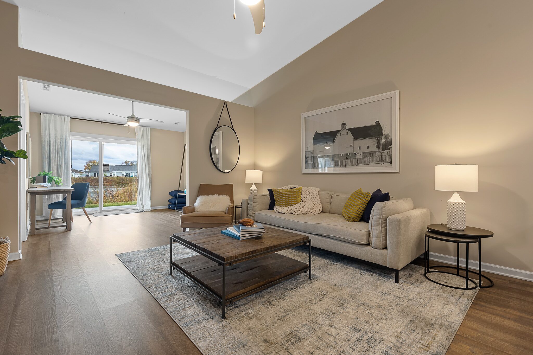Modern living room with beige walls, neutral furniture, a round mirror, framed artwork, and a coffee table on a rug; sliding glass doors lead to a dining area with outside view.