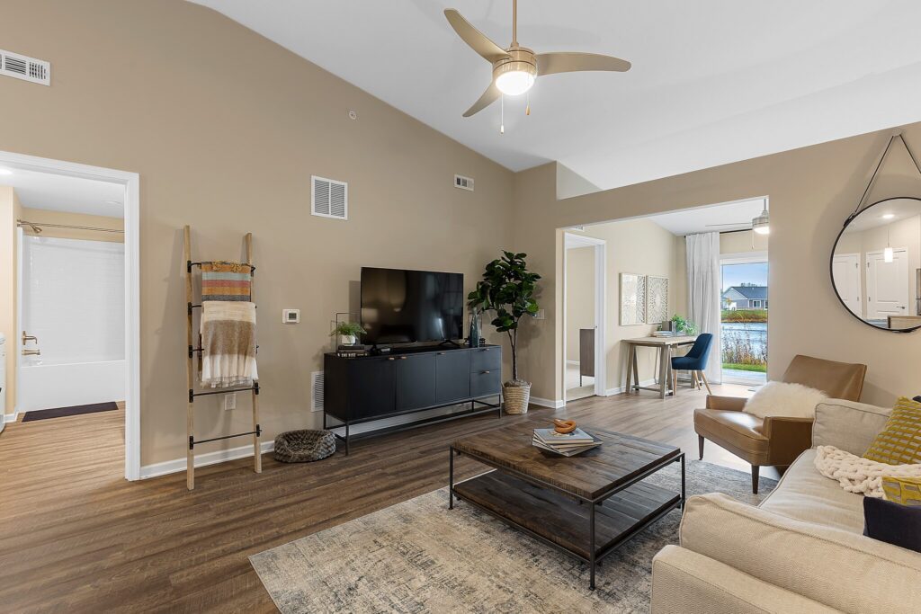 Modern living room with beige walls, wood flooring, a sofa, coffee table, TV on a black stand, a ceiling fan, and decor accents; dining area visible in the background.