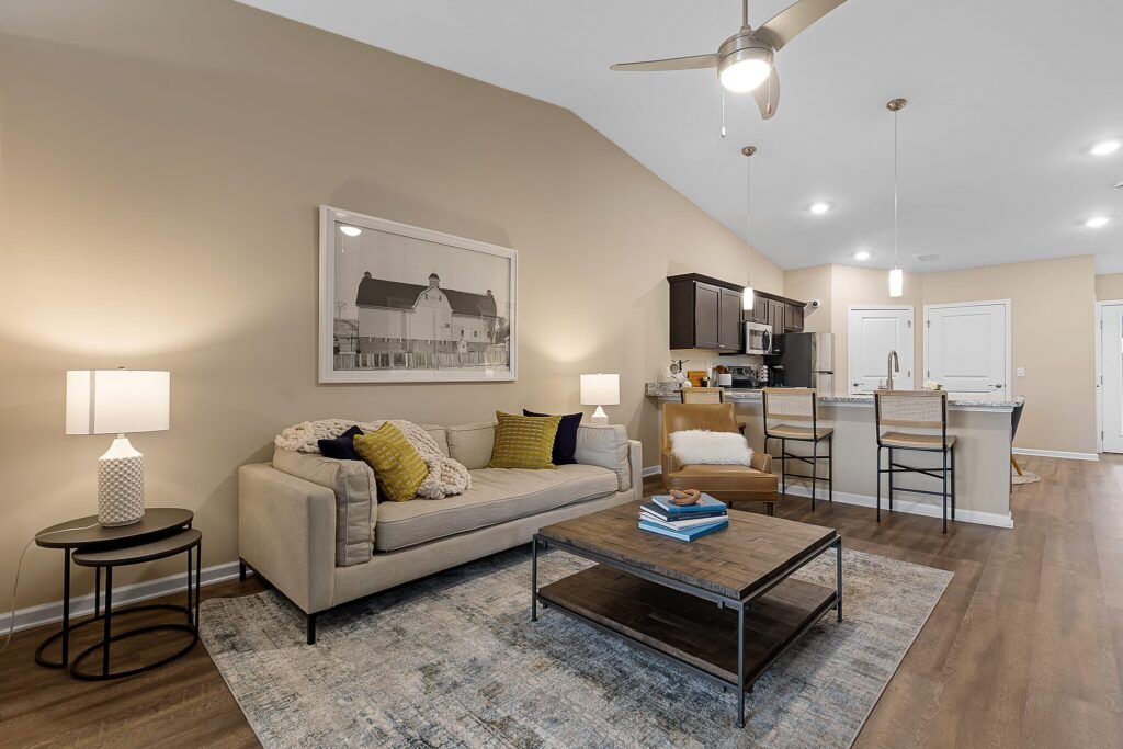 Modern living room with beige walls, a sofa, armchair, coffee table, and kitchen area with bar stools in the background. Ceiling fan and recessed lighting are visible.