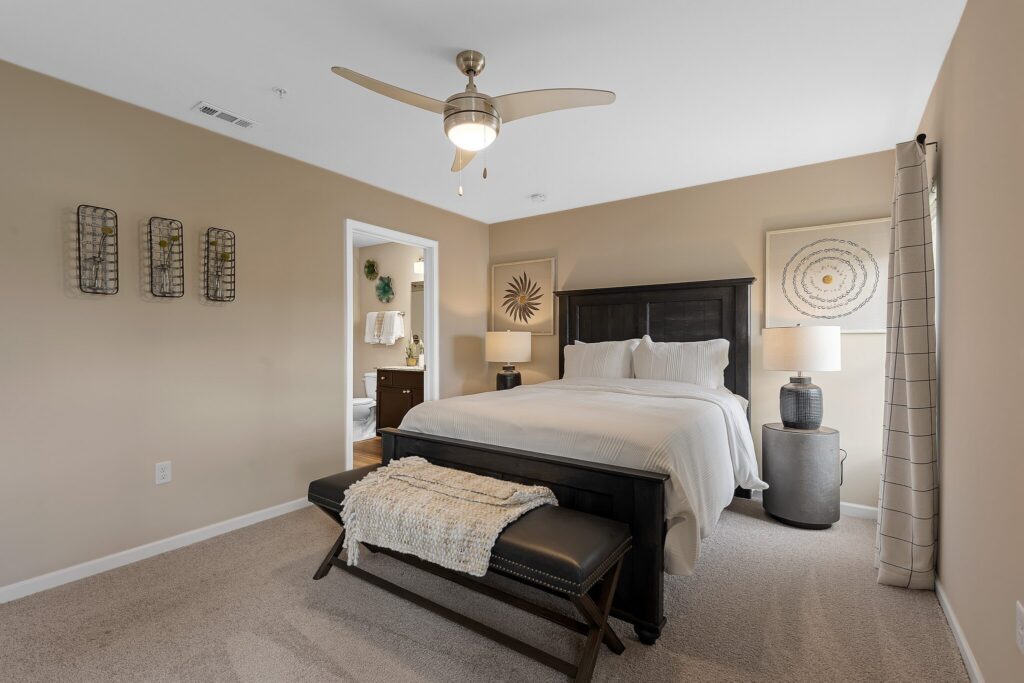 A modern bedroom with a dark wood bed frame, white bedding, two bedside lamps, a bench at the foot of the bed, and beige walls decorated with simple art.