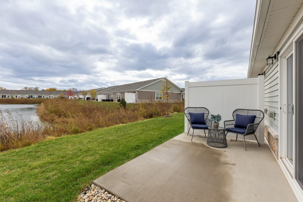 A concrete patio with two blue cushioned chairs and a small table overlooks a grassy area and a pond, with residential buildings in the background under a cloudy sky.