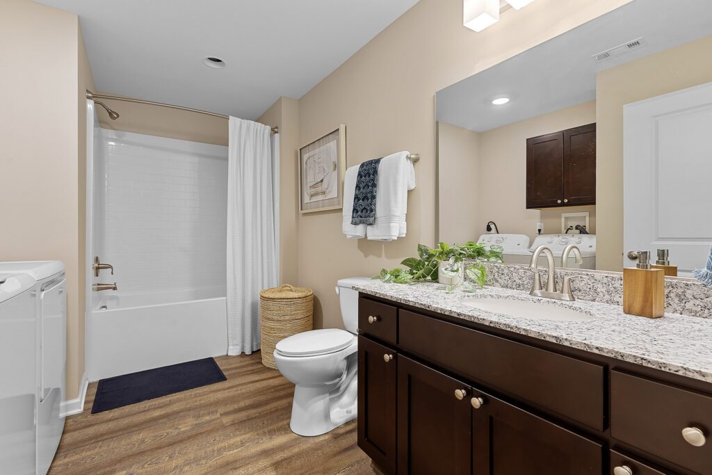 Modern bathroom with a granite countertop, dark wood cabinets, white toilet, bathtub with shower curtain, washer, and decorative plants and towels.
