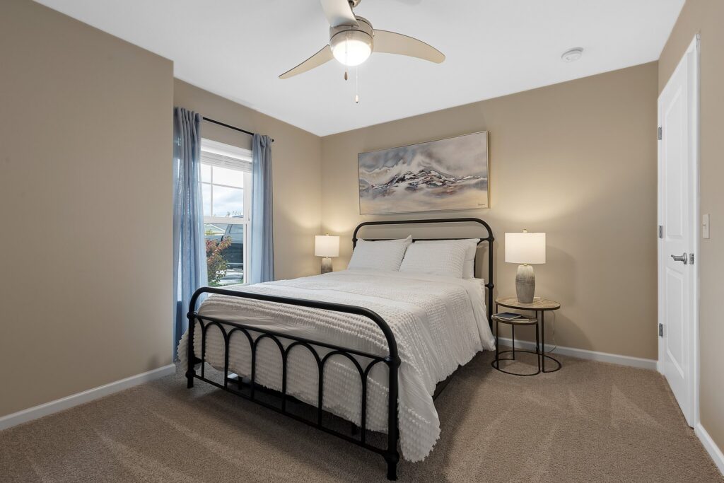 A bedroom with a metal bed frame, white bedding, two bedside tables with lamps, a large painting above the bed, beige walls, carpeted floor, and a ceiling fan.