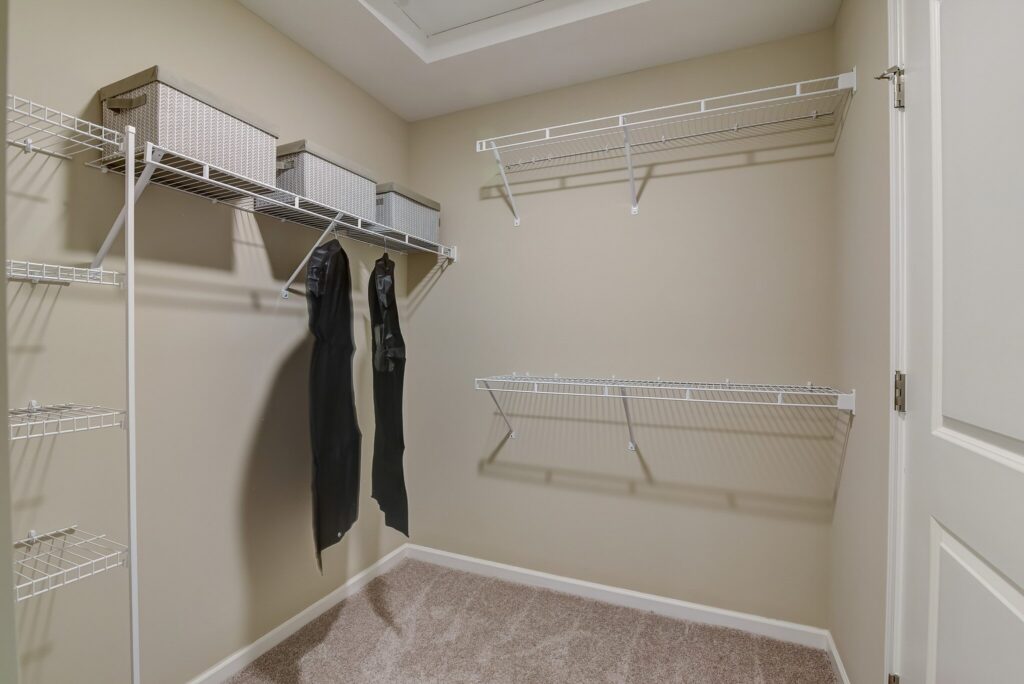 Empty walk-in closet with beige walls, carpeted floor, wire shelving on three sides, two black hanging garments, and three storage baskets on the top shelf.