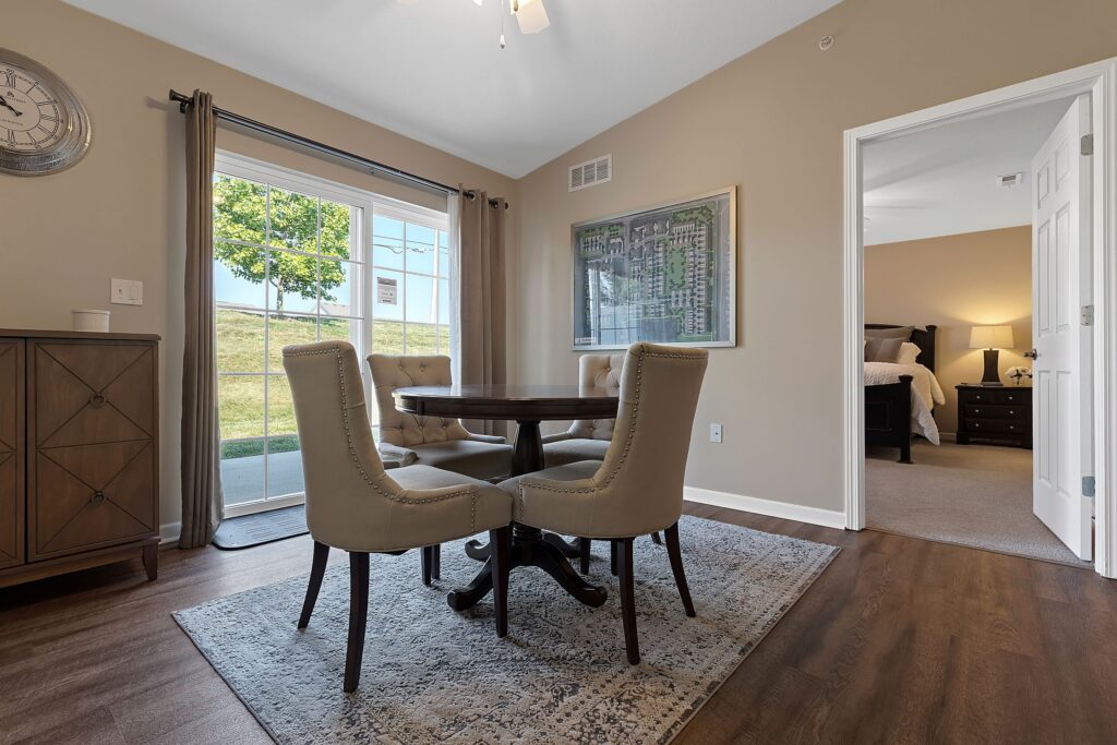 A dining area with a round table and four upholstered chairs, a rug underneath, and a view into a bedroom; large window with outdoor view on the left.
