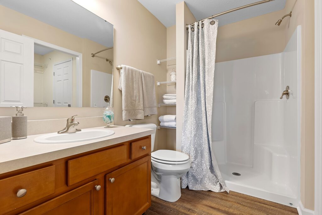 Bathroom with wooden cabinets, a countertop sink, toilet, towel rack, and a walk-in shower with a patterned curtain and shelving with neatly folded towels.