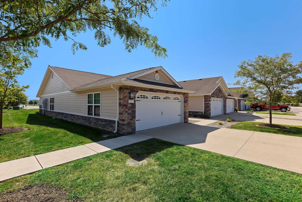 Single-story suburban house with a two-car garage, brick and siding exterior, concrete driveway, and a well-maintained lawn on a clear day.