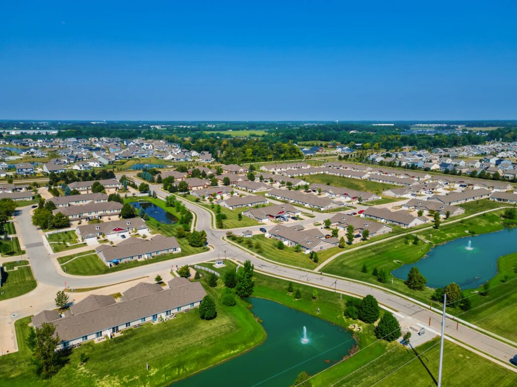 Aerial view of a suburban neighborhood with houses, roads, green lawns, ponds, and fountains under a clear blue sky.