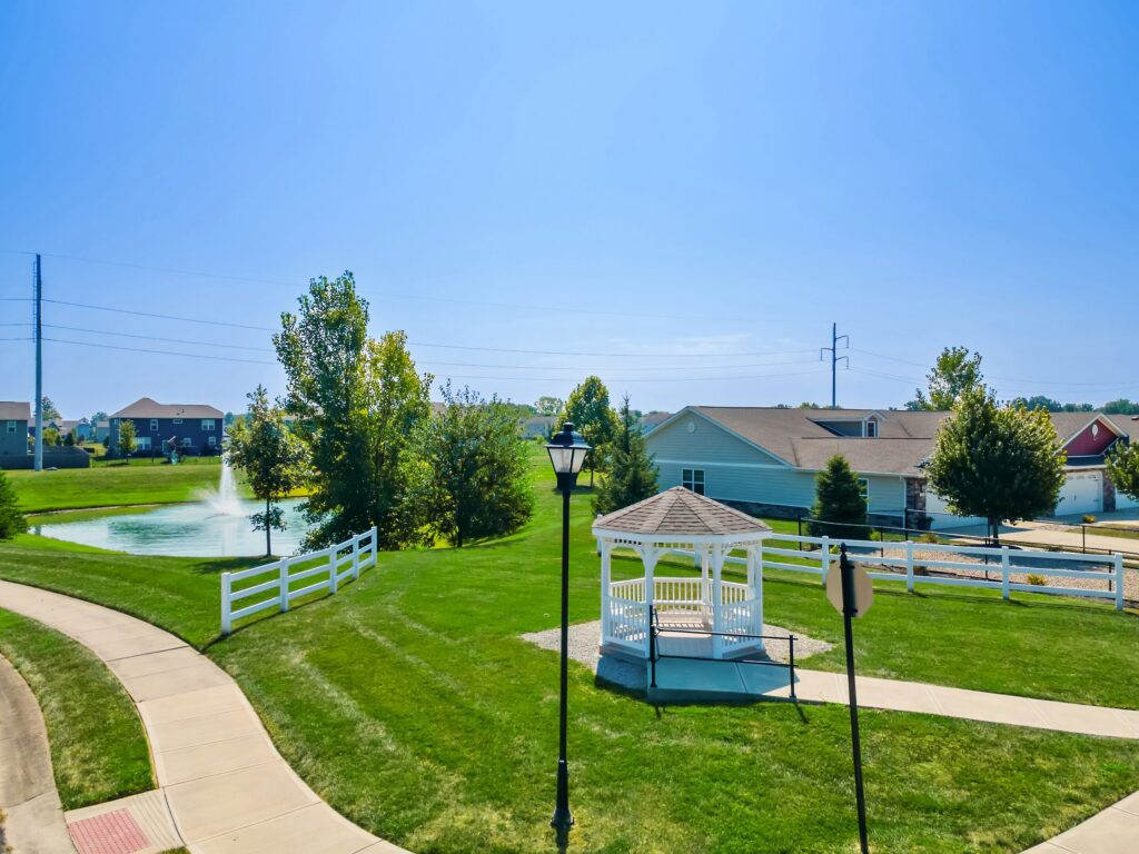 A white gazebo sits on a lawn near a sidewalk, with a pond, fountain, houses, and trees in the background under a clear blue sky.