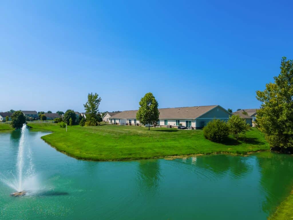 A small pond with a fountain, surrounded by green grass, trees, and single-story houses under a clear blue sky.