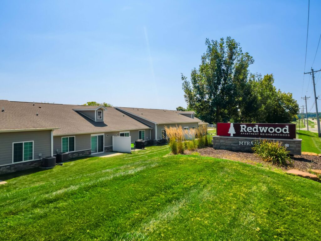 Single-story apartment buildings with gray siding next to a landscaped lawn and a sign reading "Redwood Apartment Neighborhoods" and "Myra Ct." under a blue sky.