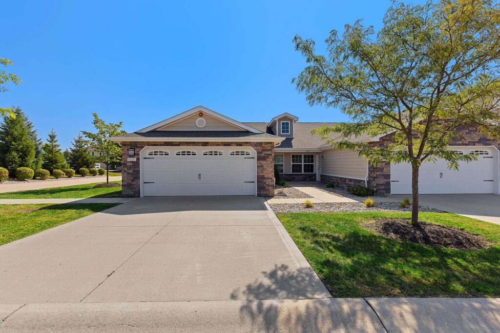 A single-story suburban duplex with stone and siding exterior, two white garage doors, a driveway, and a tree in the front yard under a clear blue sky.