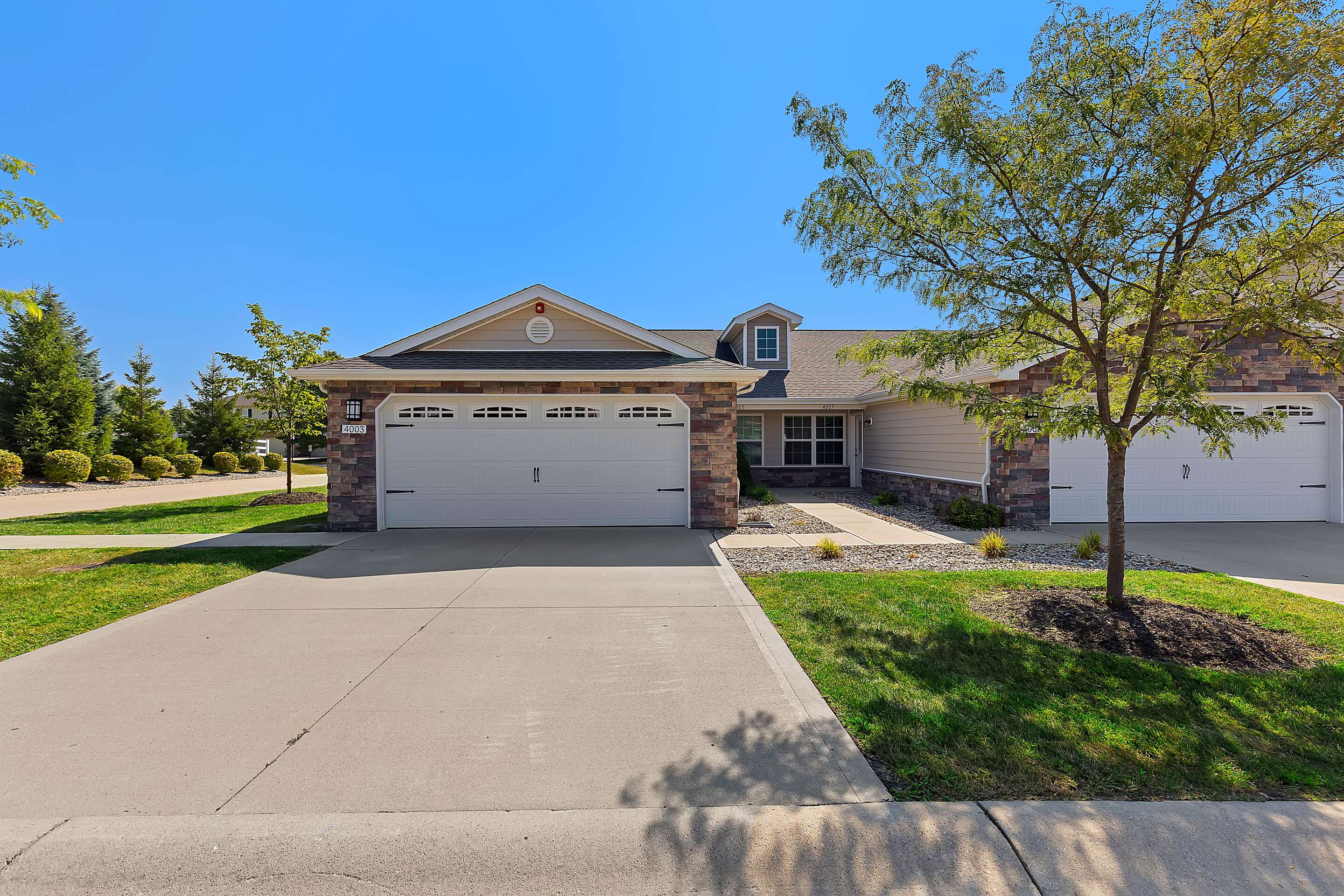 A single-story suburban duplex with stone and siding exterior, two white garage doors, a driveway, and a tree in the front yard under a clear blue sky.