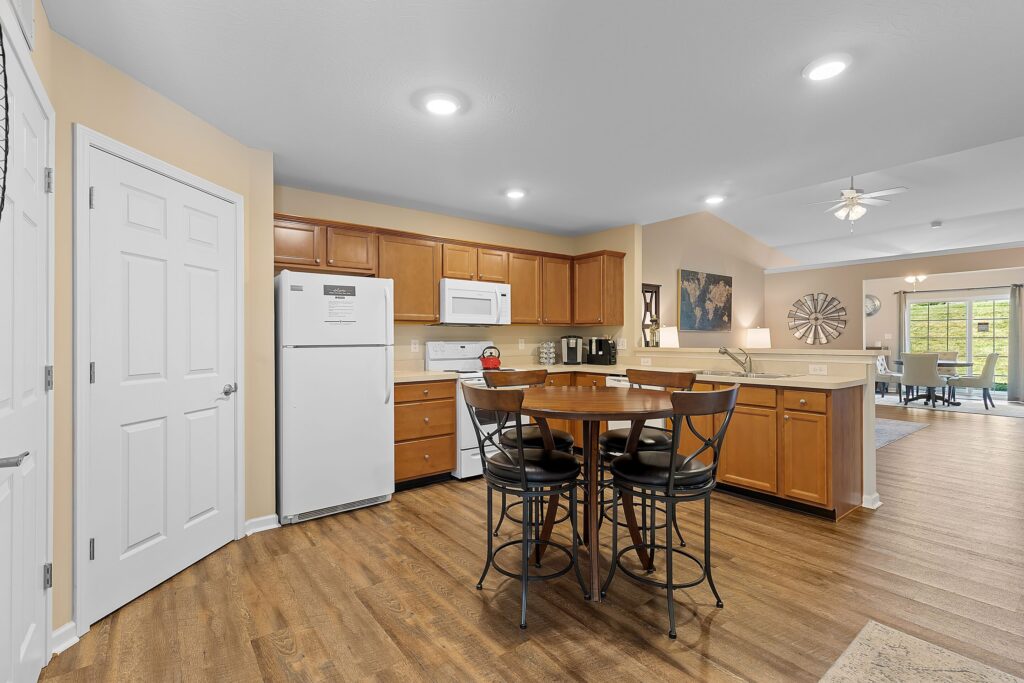 Kitchen with wooden cabinets, white appliances, round dining table with four chairs, and a view into a living area with large windows.