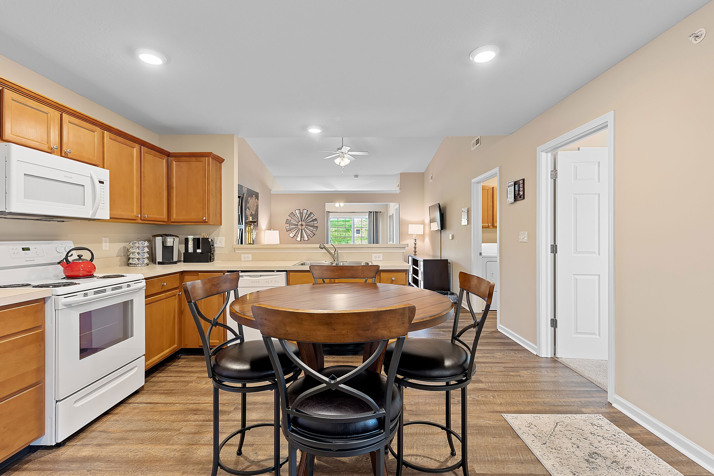 A kitchen with wooden cabinets, white appliances, a round dining table with four chairs, and an open view into a living area.