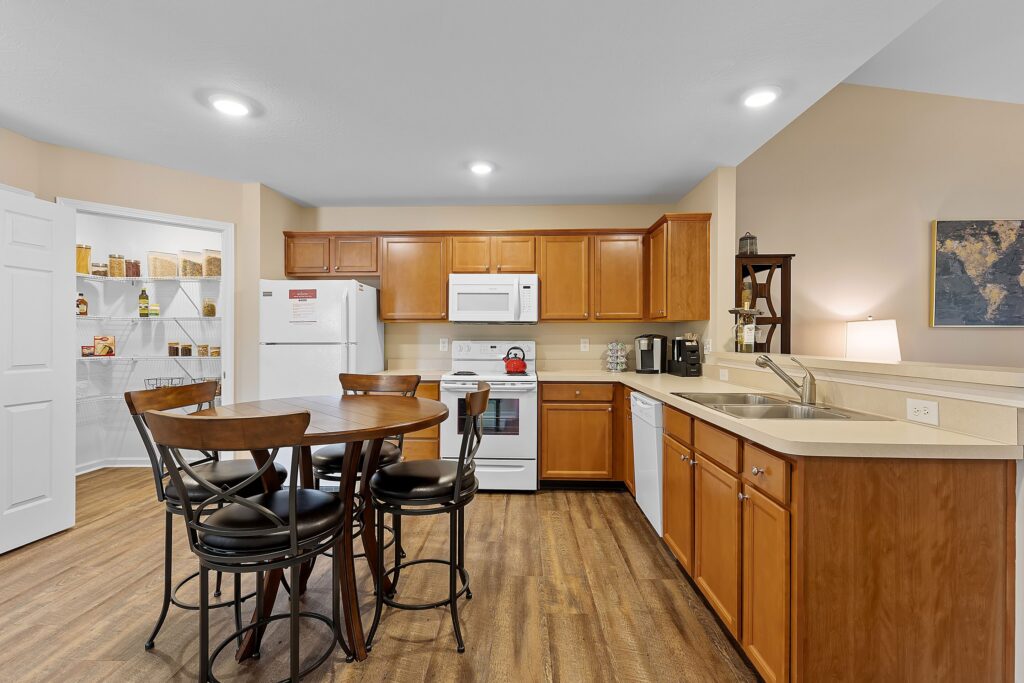Modern kitchen with wooden cabinets, white appliances, round dining table with four chairs, and adjacent pantry with shelves holding food containers.