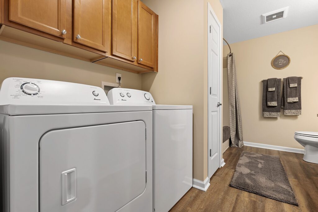 Laundry room with a white washer and dryer, wooden cabinets above, and view into a bathroom area with towels, toilet, rugs, and shower curtain.