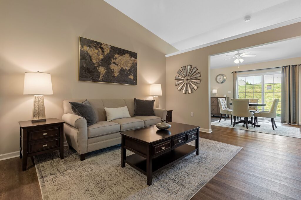 A living room with a beige sofa, two side tables with lamps, a coffee table, wall art, and an adjacent dining area with a table and chairs near large windows.