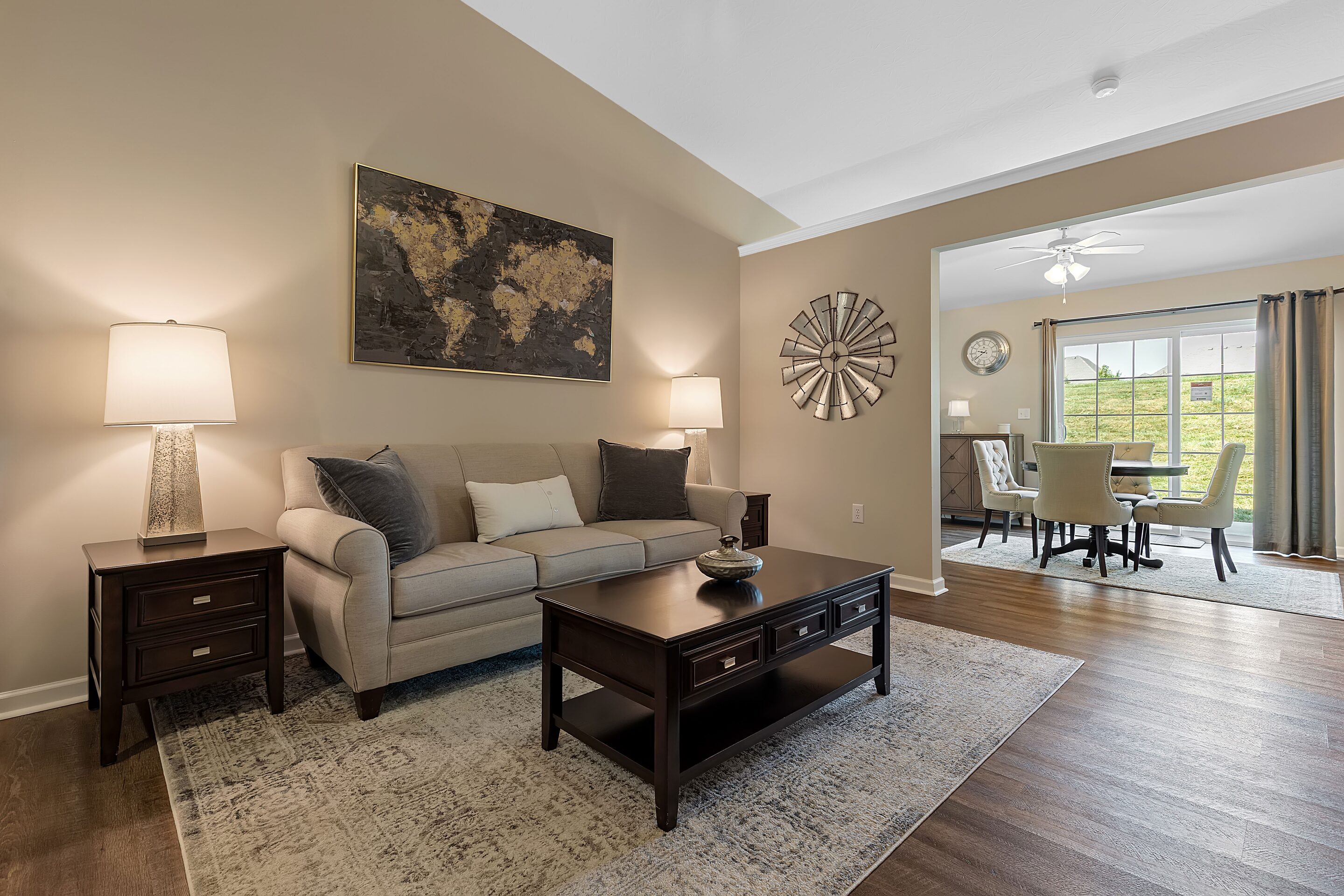 A living room with a beige sofa, two side tables with lamps, a coffee table, wall art, and an adjacent dining area with a table and chairs near large windows.