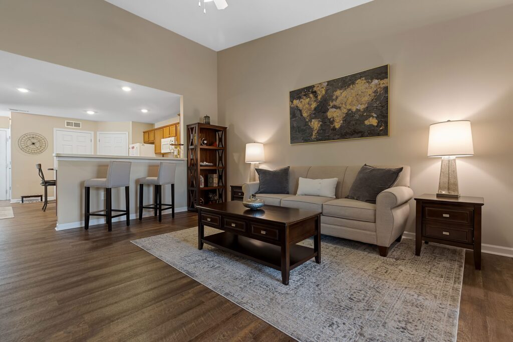 A modern living room with a beige sofa, two lamps, a wooden coffee table, and an area rug, adjacent to a kitchen with a breakfast bar and three stools.