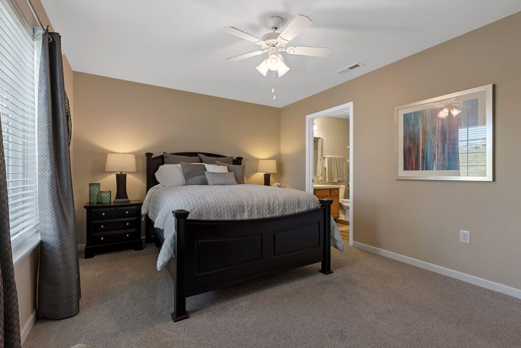 Bedroom with a dark wood bed, two nightstands with lamps, neutral walls, carpeted floor, large window with curtains, and a door leading to an adjoining bathroom.