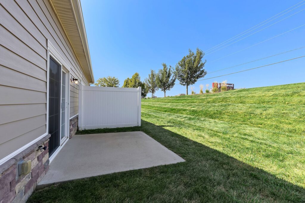 A small concrete patio next to a beige house with sliding glass doors, adjacent to a white privacy fence, overlooking a grassy yard and trees under a clear blue sky.