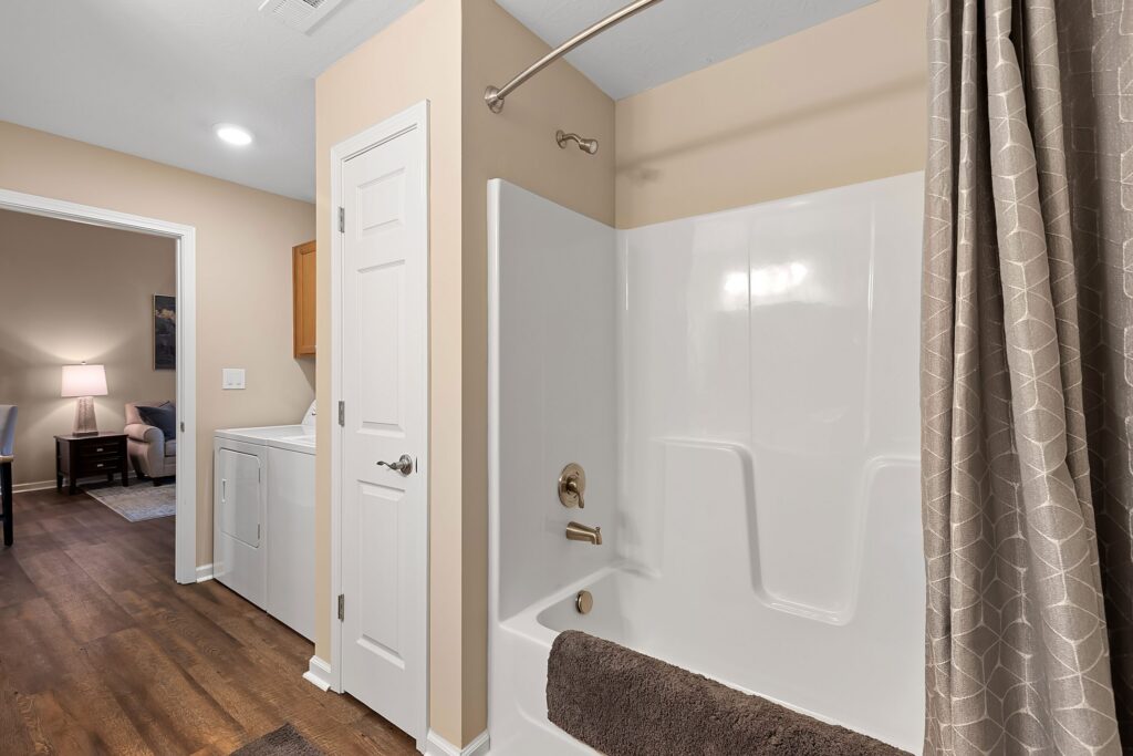 A bathroom with a white bathtub and shower, a brown towel draped over the tub, beige walls, and a view into a laundry area with a washer and dryer.