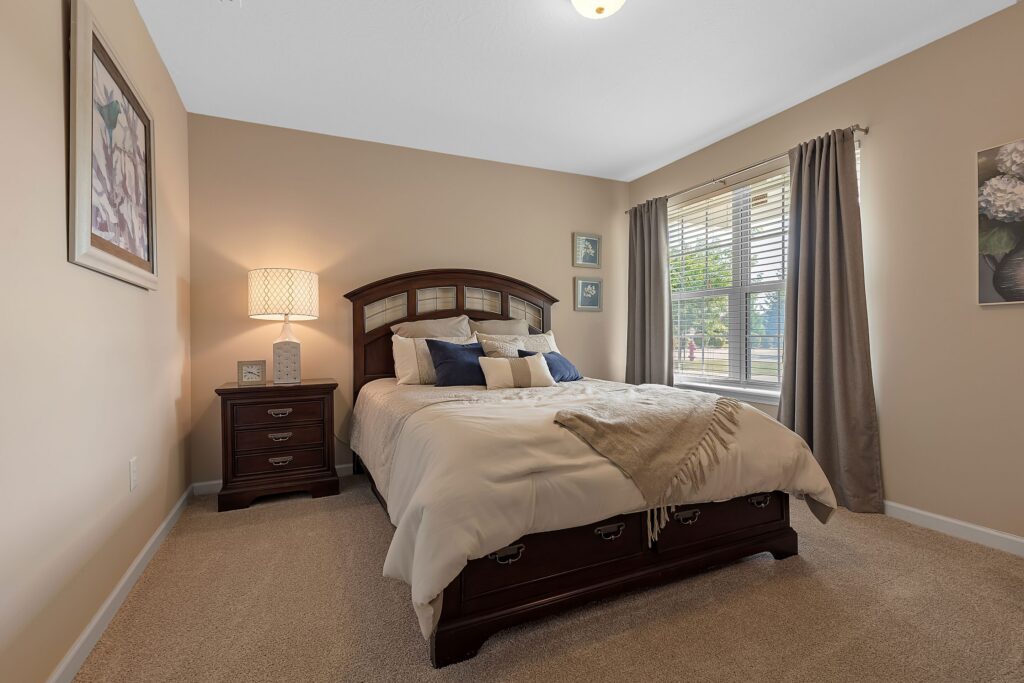 A neatly made bed with beige bedding and blue accent pillows is centered in a carpeted bedroom, with a wooden nightstand, lamp, wall art, and a window with curtains.
