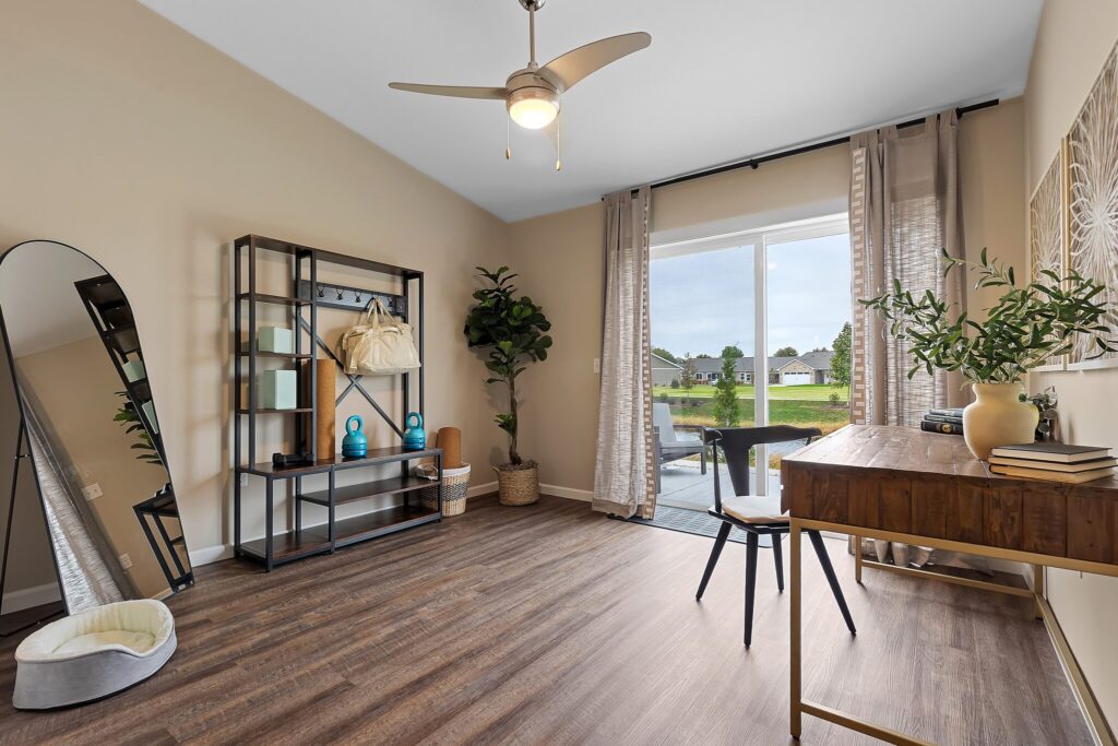 Modern home office with a wooden desk, black chair, bookshelf, large mirror, potted plants, and a dog bed near a sliding glass door overlooking a backyard.