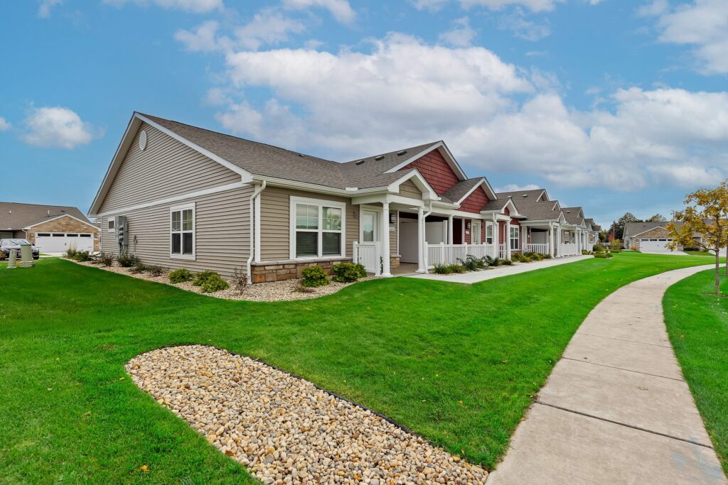 Single-story suburban houses with front porches and manicured lawns line a sidewalk under a partly cloudy sky.