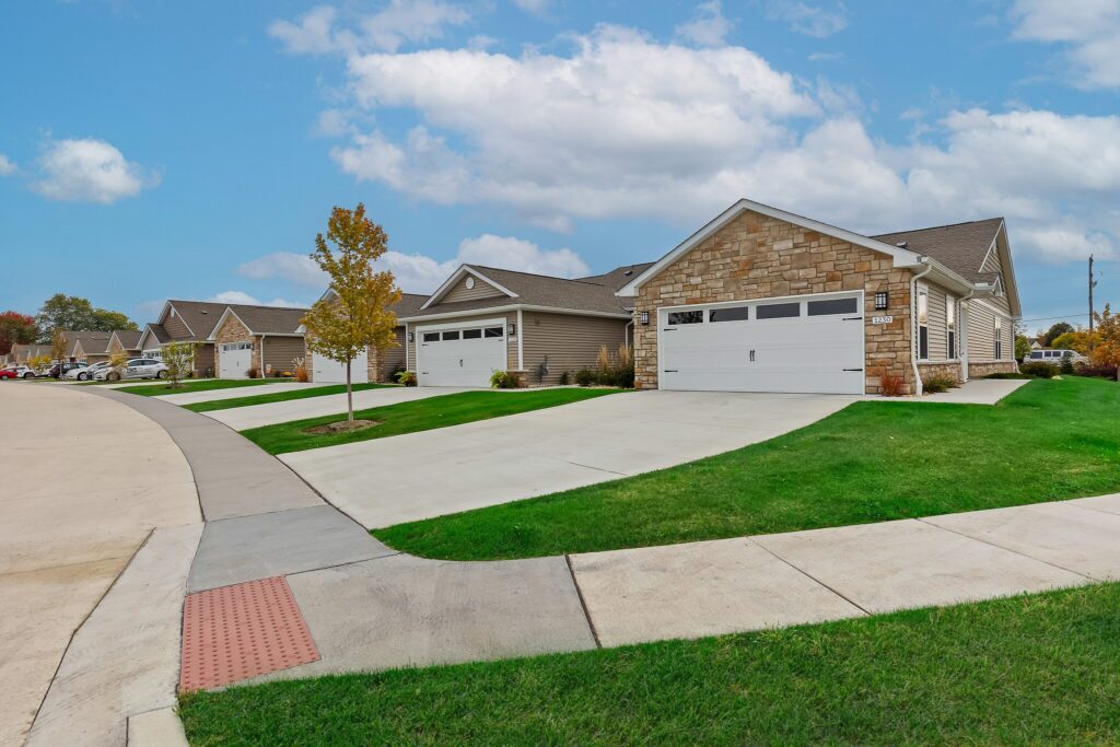 Row of modern single-story suburban houses with attached garages, neatly maintained lawns, and a wide concrete sidewalk under a partly cloudy sky.