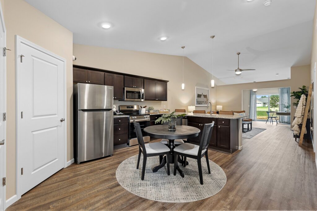 Modern open-concept kitchen and dining area with dark cabinets, stainless steel appliances, round dining table, and view into living room with large windows.